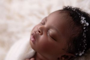 Sleeping baby wearing delicate pearl headpiece rests peacefully against soft cream-colored background.