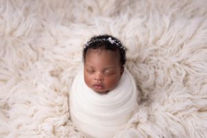 A sleeping newborn baby wrapped in white fabric rests peacefully on soft cream-colored fur bedding.