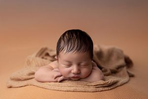 A peaceful sleeping newborn baby wrapped in soft beige fabric against a warm neutral background.
