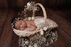 Sleeping newborn baby nestled in a rustic basket surrounded by delicate white baby's breath flowers on wooden floor.