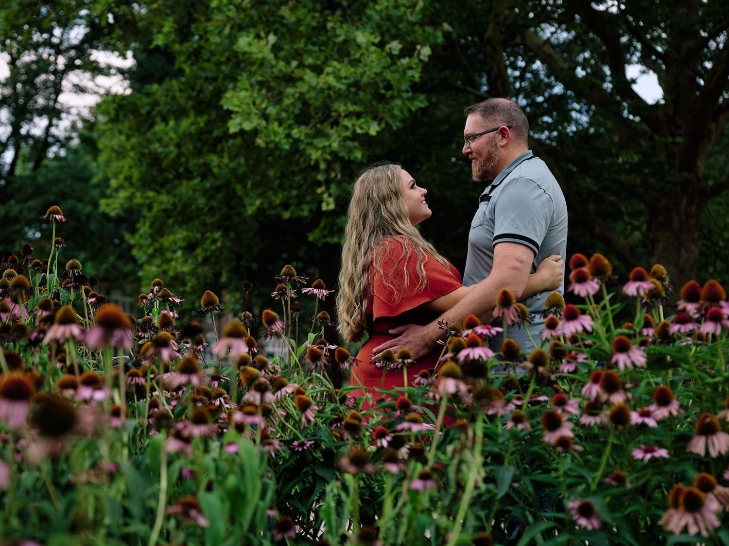 A romantic couple embraces on a wooden footbridge surrounded by lush garden greenery and hanging vines.