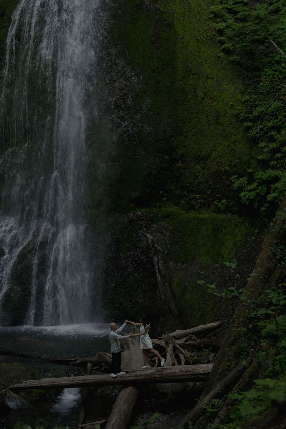 Two people stand on fallen logs near a majestic waterfall in a lush forest setting Marymere falls in Olympic National Park..
