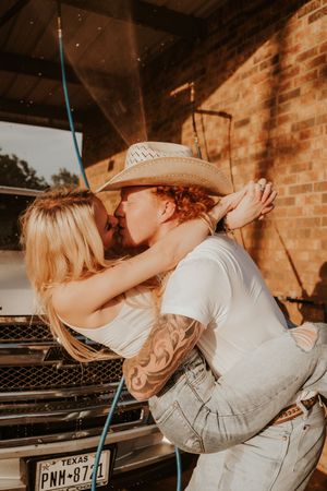 Romantic couple sharing kisses against a rustic brick wall backdrop at sunset.