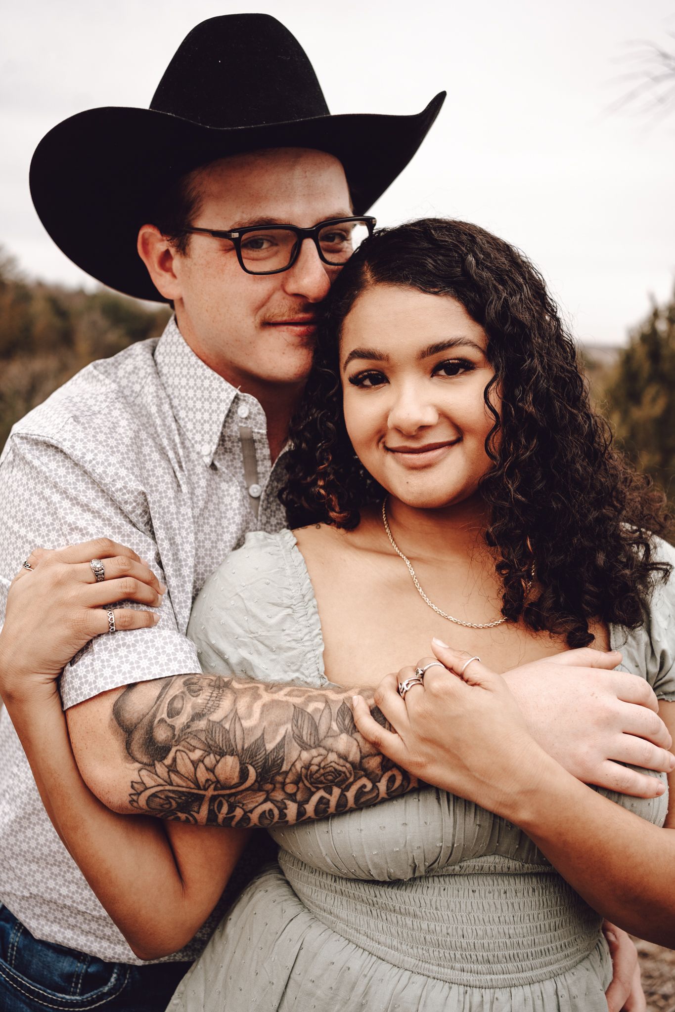 Romantic couple embraces in casual western attire against natural outdoor backdrop.