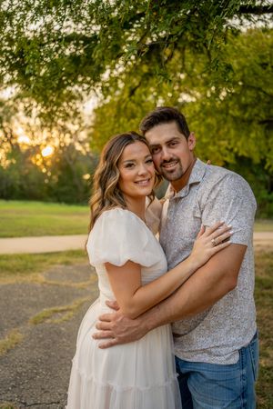 A couple embraces lovingly in a park during golden hour with trees in the background.