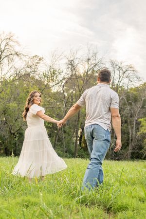 A couple dances joyfully through a green meadow with trees in the background.