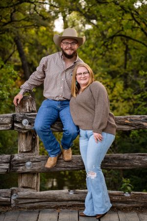 Couple sits together on rustic wooden fence in countryside setting.