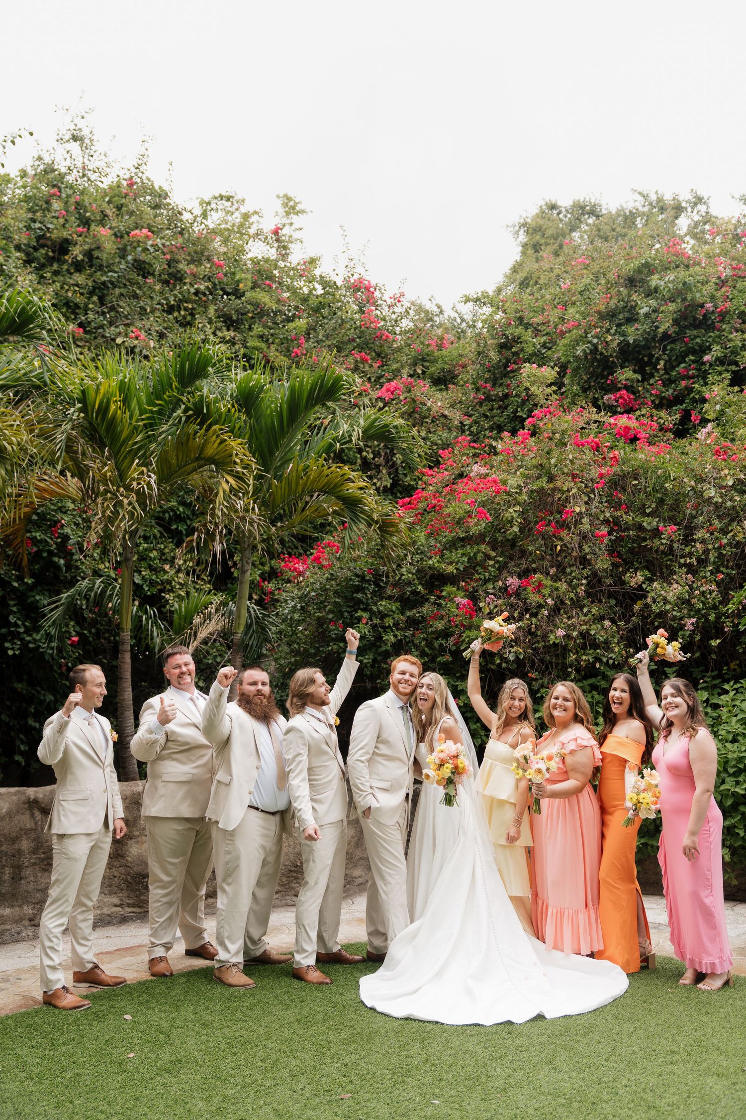 Wedding party in pastel colors poses together on a lawn with blooming bougainvillea and palm trees in the background.