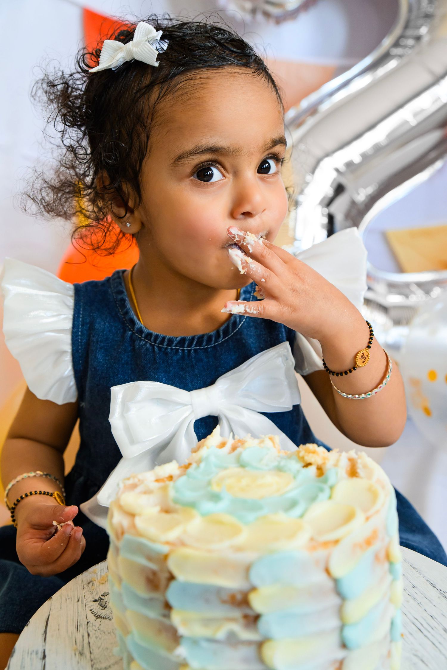 Young girl in denim dress with white bow licking frosting off her fingers beside a pastel blue birthday cake.