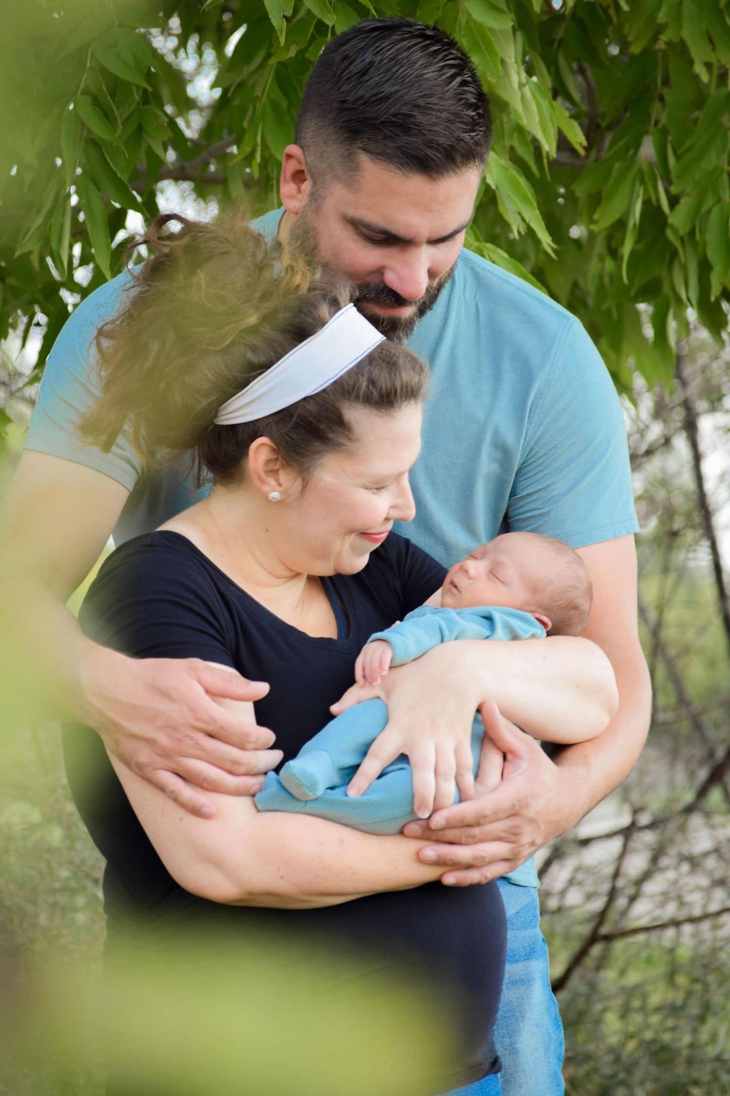 Baby in light blue outfit being held tenderly outdoors among green foliage.
