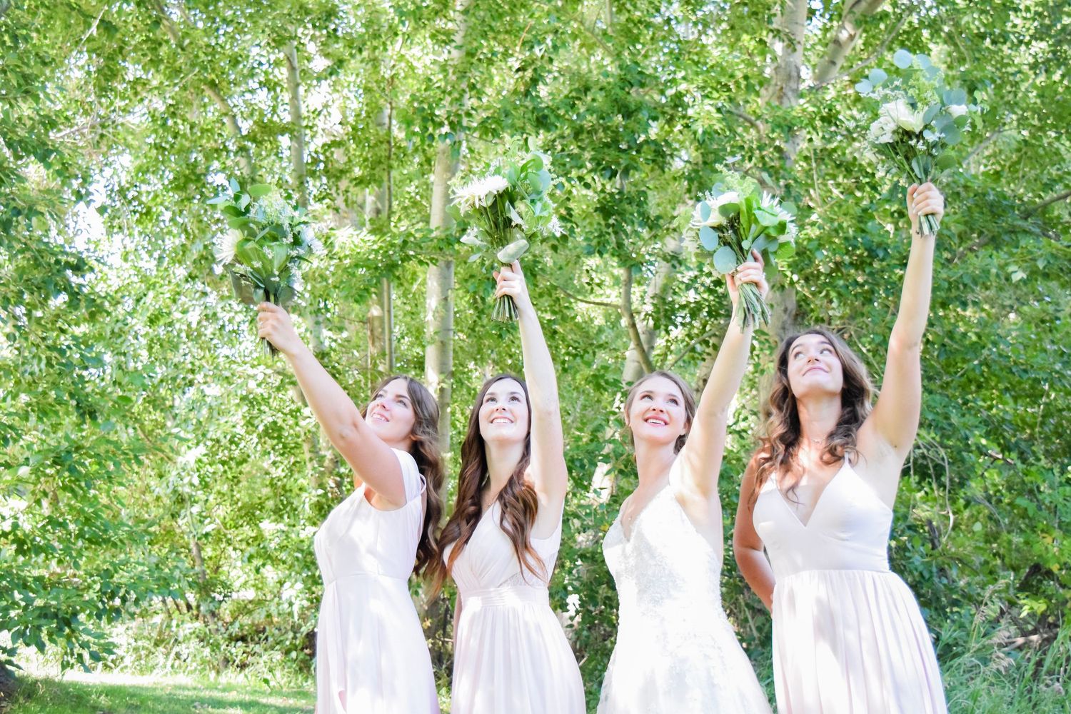 Bridesmaids in white dresses celebrate with arms raised against green foliage background.