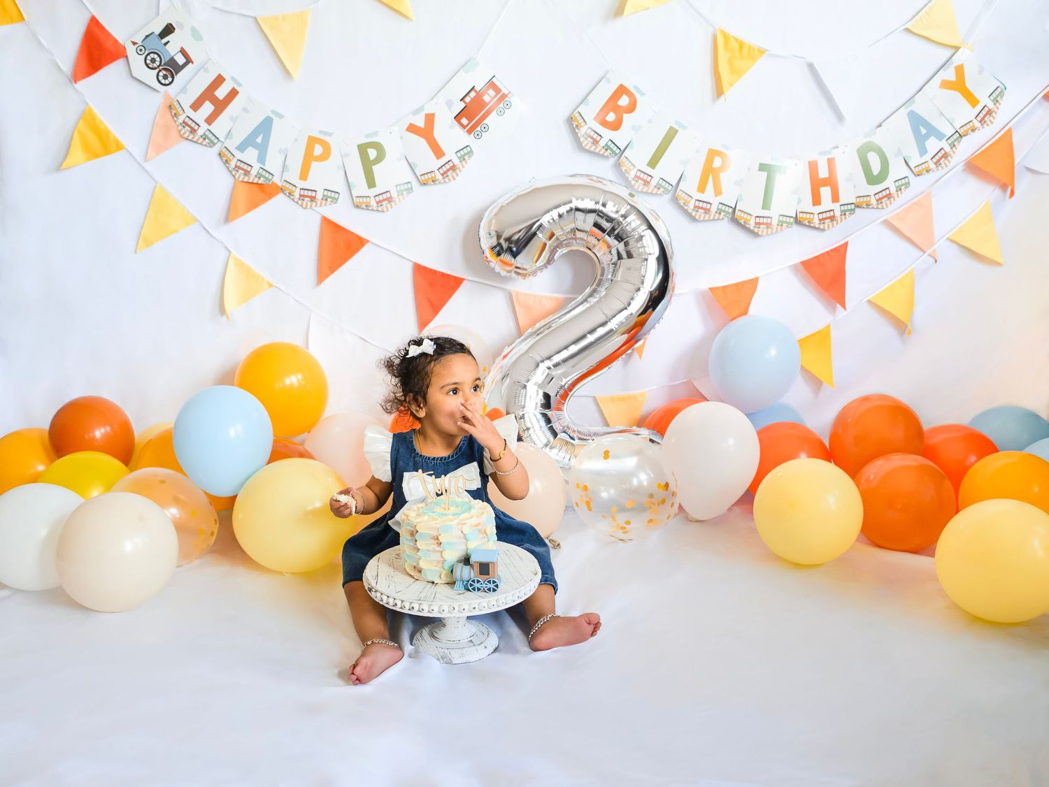 Toddler girl celebrating 2nd birthday with cake smash, surrounded by colorful balloons and Happy Birthday banner.