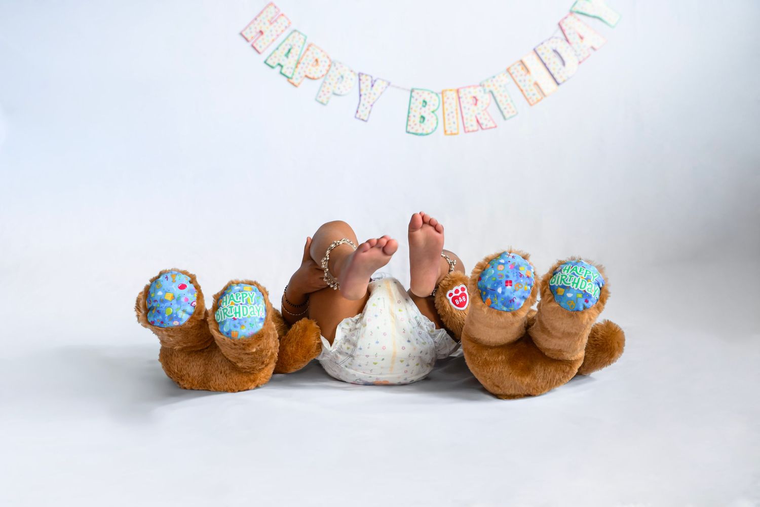 Baby feet with colorful cupcake decorations beside teddy bears under a Happy Birthday banner on white background.