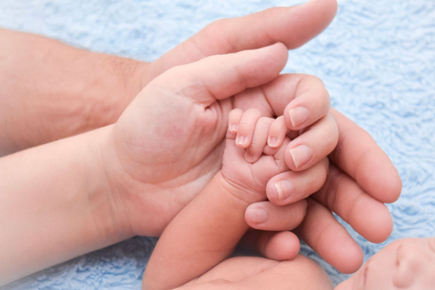 Close-up of gentle hands holding tiny baby fingers on soft blue fabric.