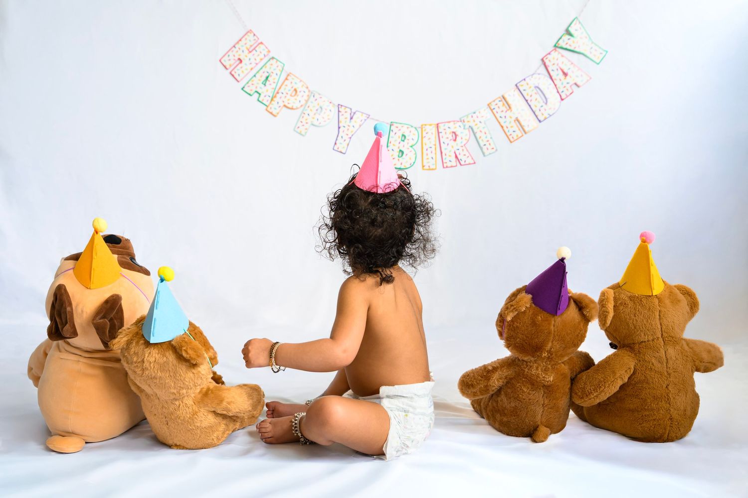 Baby in diaper sits with teddy bears wearing party hats under a Happy Birthday banner for a first birthday photo.