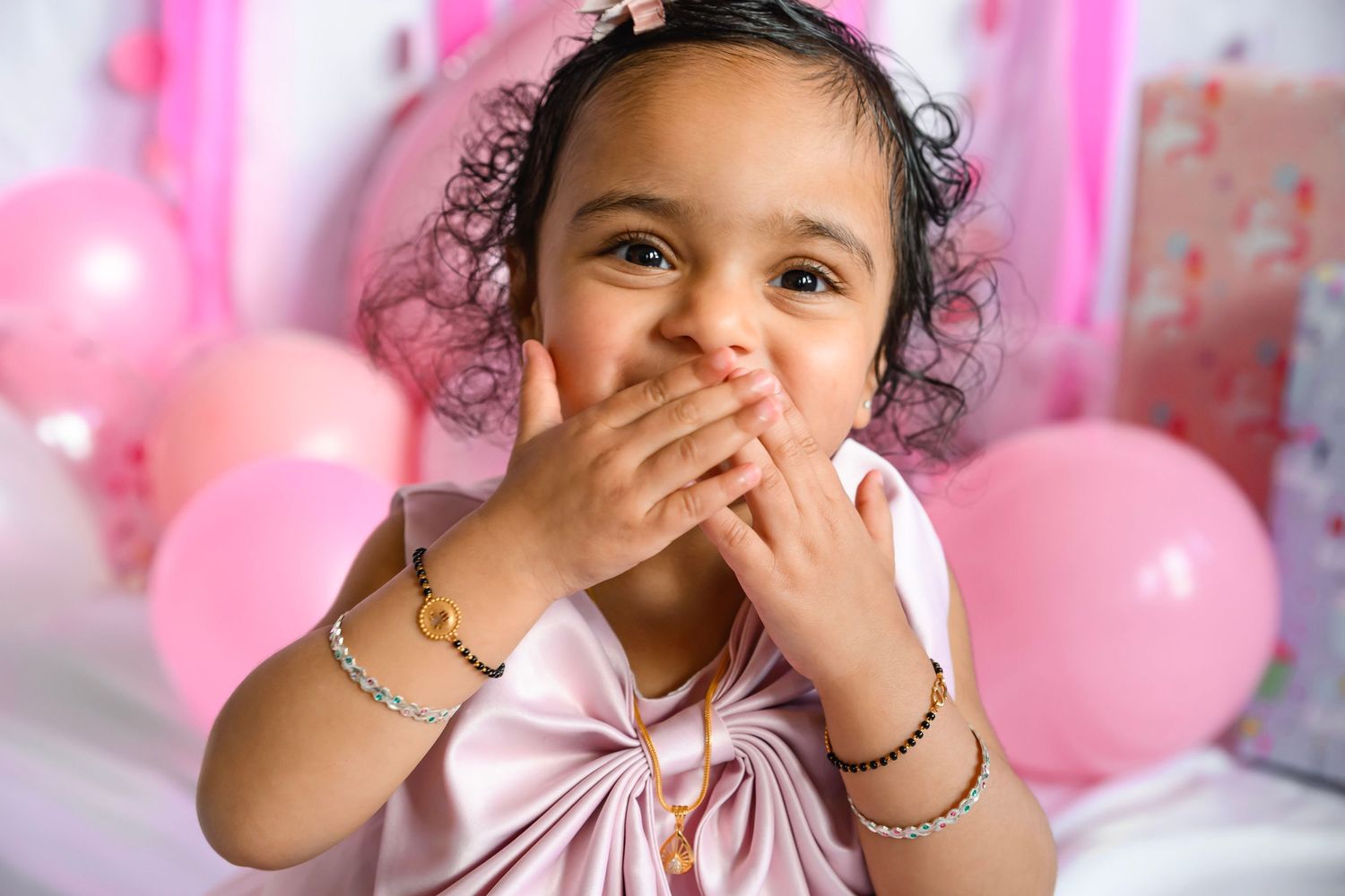 A joyful toddler girl in a pink dress covers her mouth with her hand, surrounded by pink balloons at a birthday party.