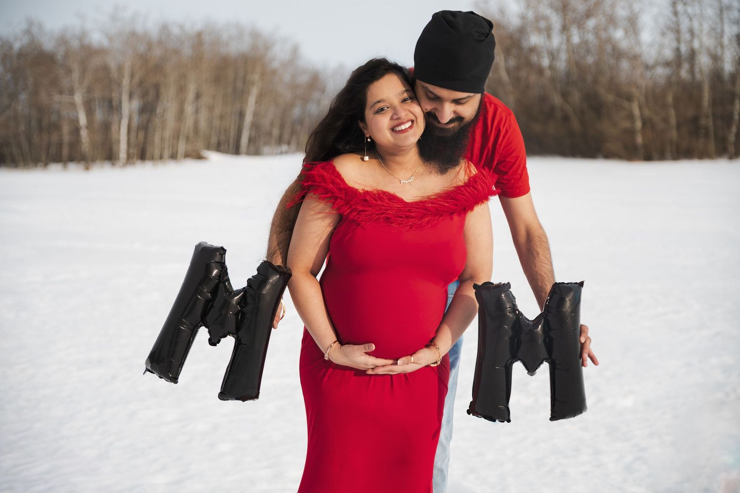 Pregnant woman in red gown with partner holding black letter balloons in snowy winter maternity photo shoot.