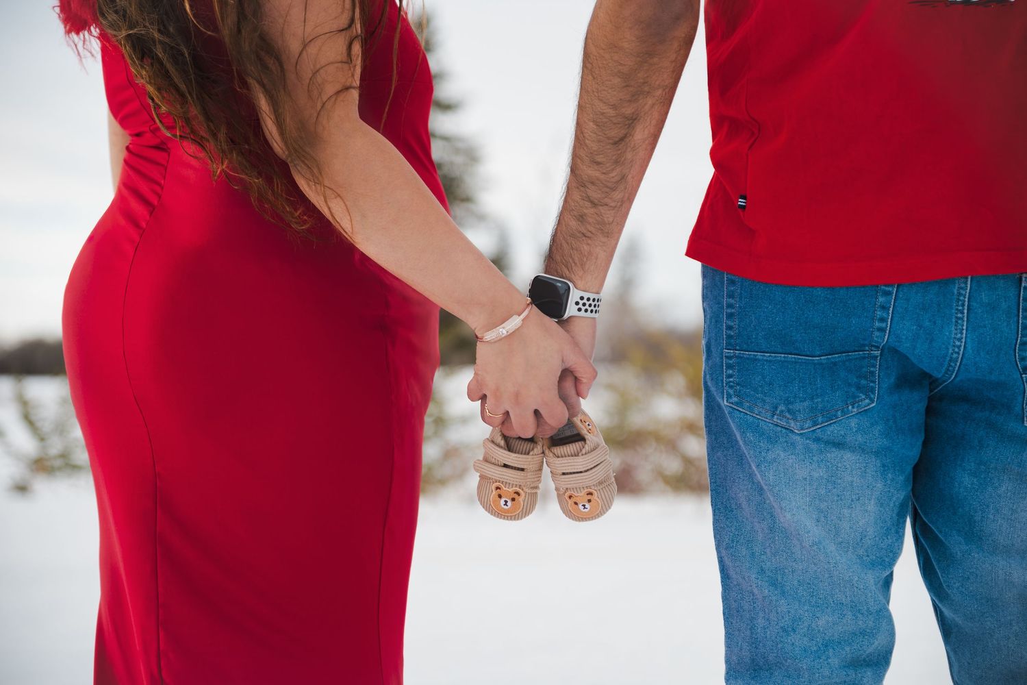 Couple in red holding tiny baby shoes in snow, announcing pregnancy in a winter maternity photo.