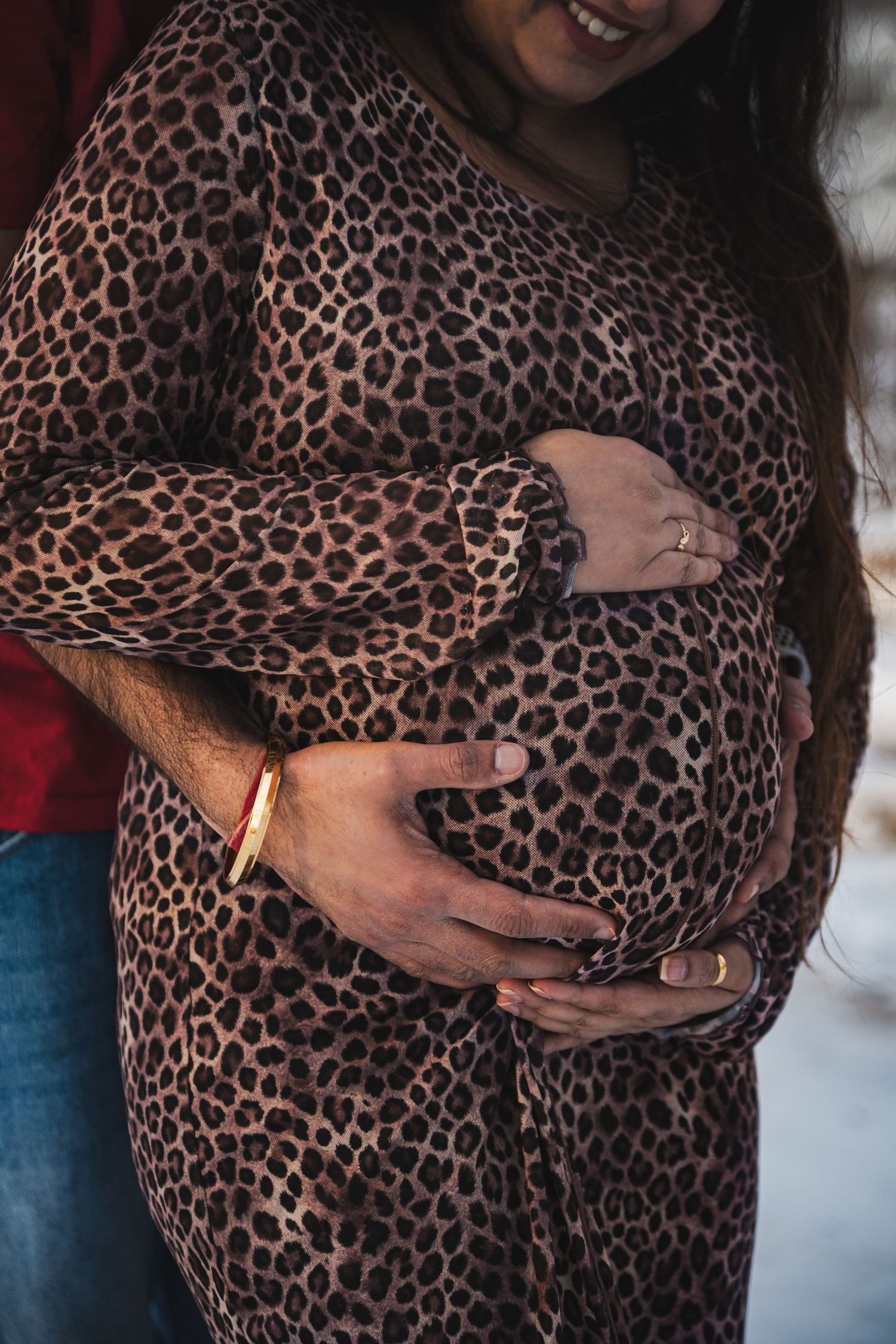 Pregnant woman in leopard print dress cradling baby bump with partner's hands around her belly.