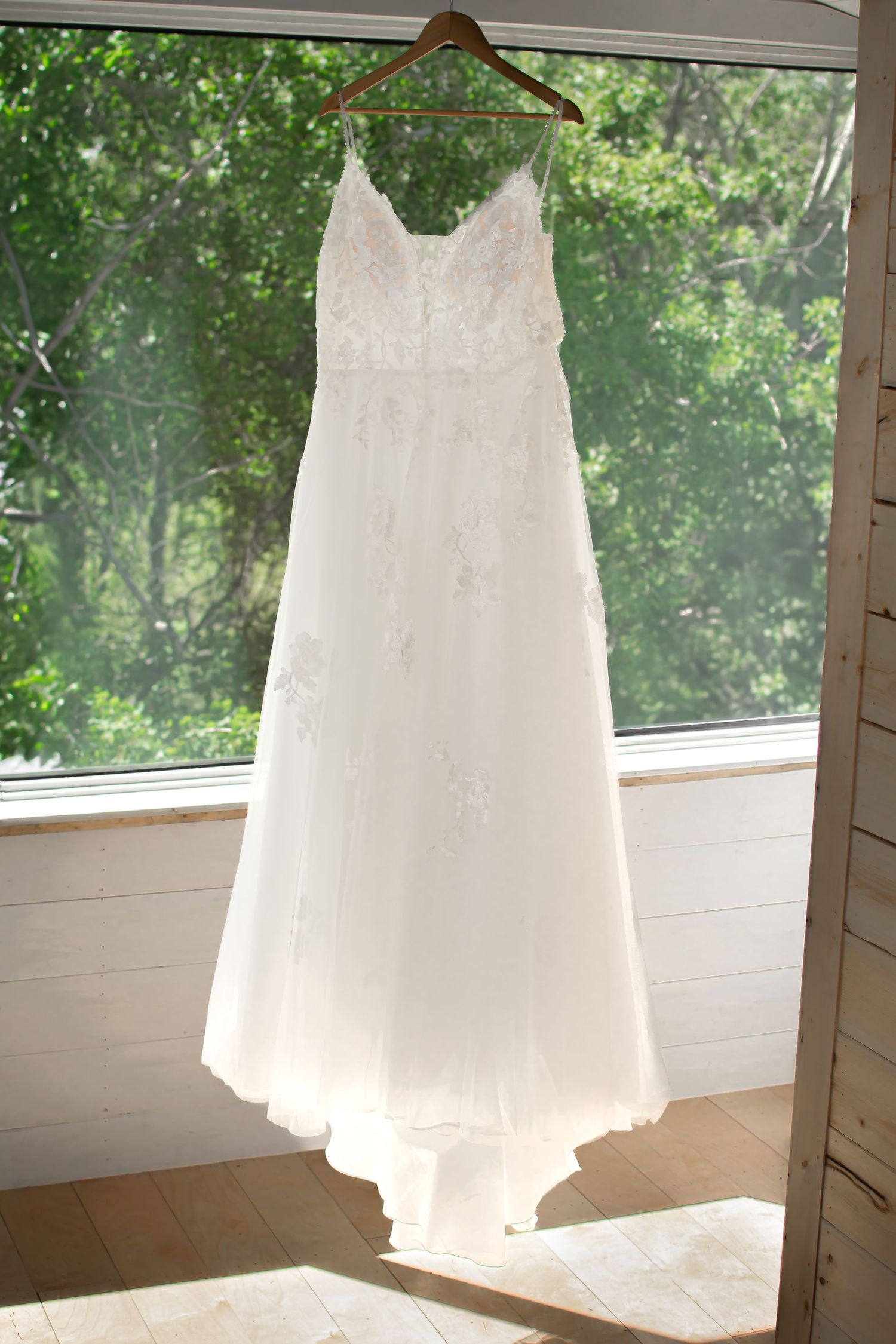 White lace wedding dresses hanging in sunlit window create ethereal silhouettes.