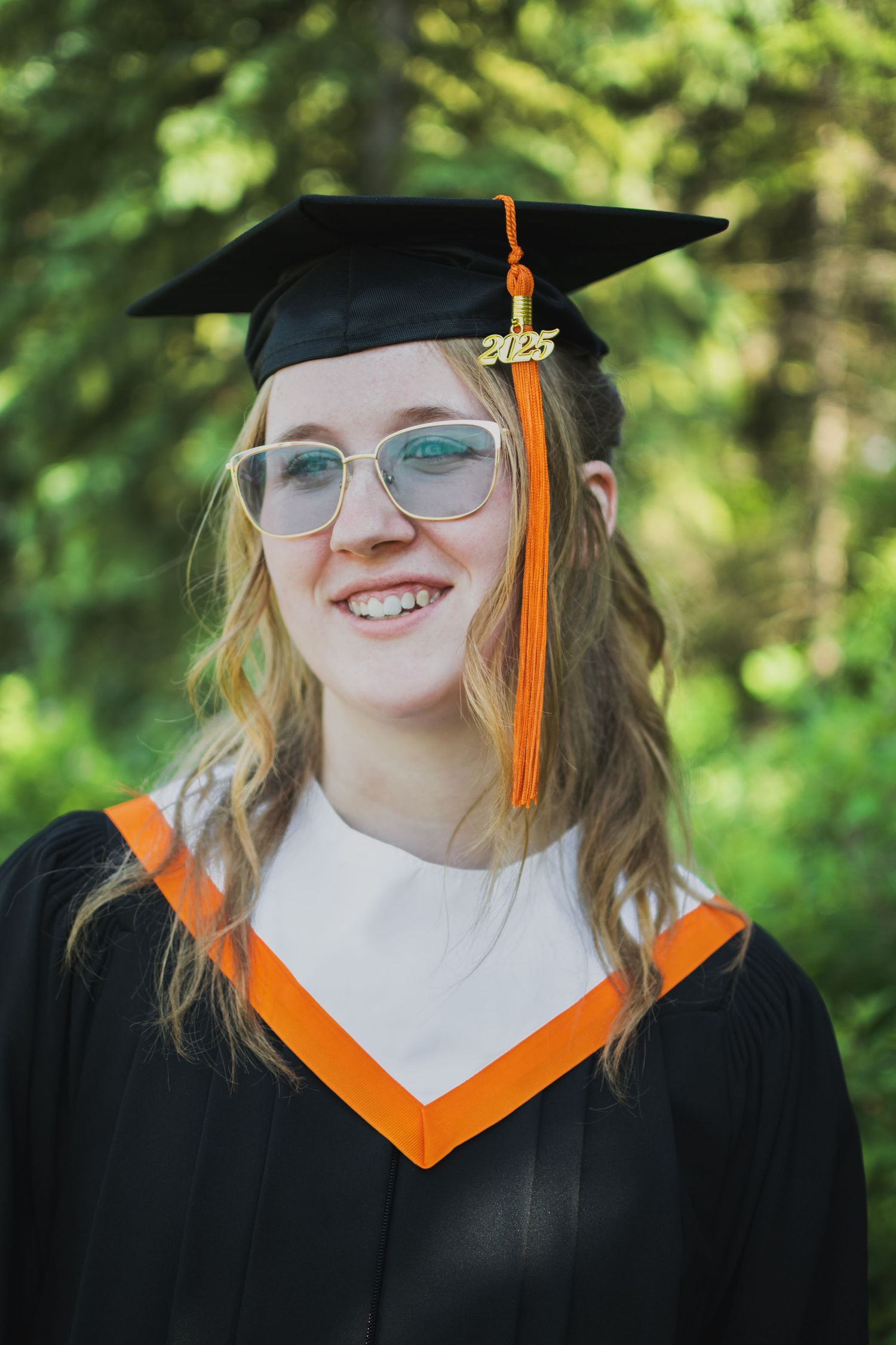 A graduate in cap and gown with orange tassel and glasses smiles during outdoor photo session.