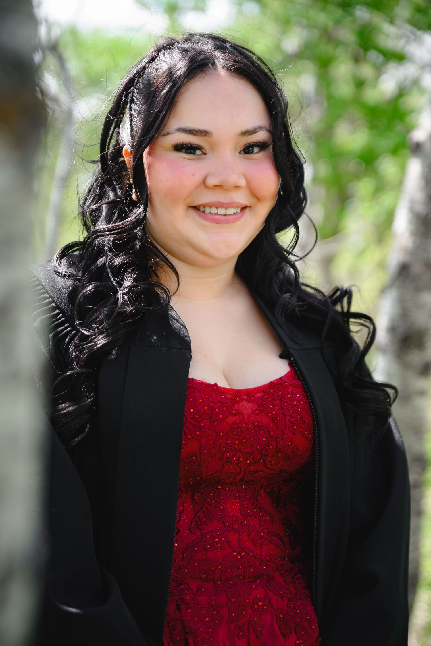 Young woman with curly black hair smiling in a red dress and black jacket outdoors.