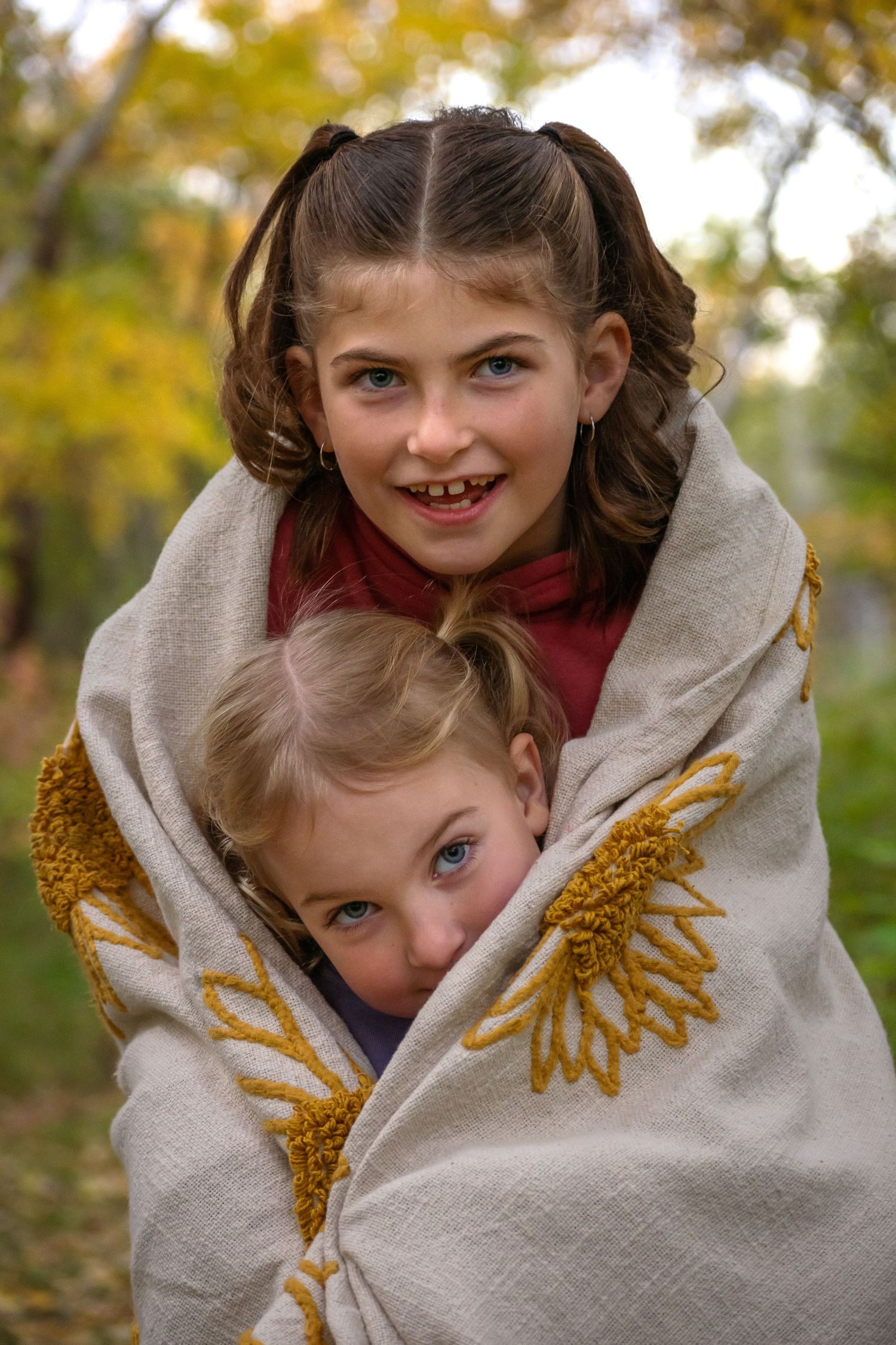 Two children share a cozy moment wrapped in a beige blanket with gold wing designs.