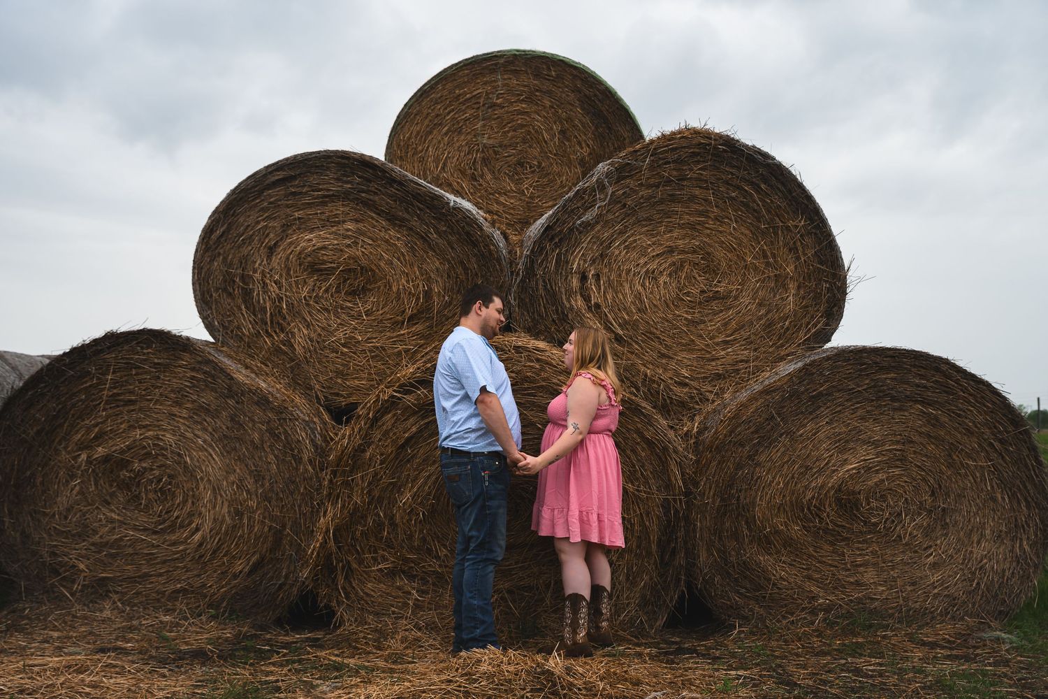 Couple holding hands in front of large round hay bales on a farm, woman in pink dress and boots.