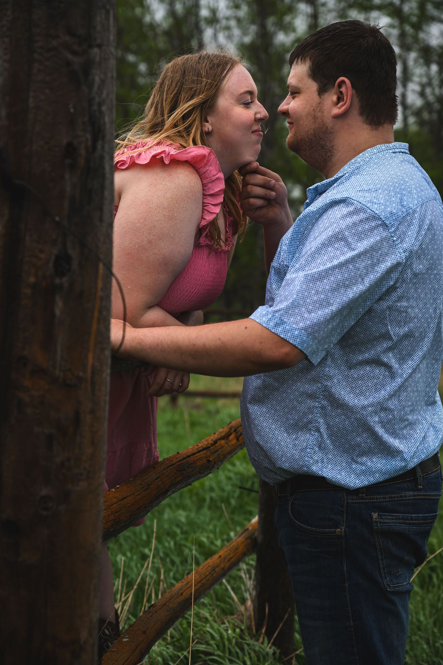A couple shares a romantic moment by a rustic wooden fence, surrounded by lush greenery.