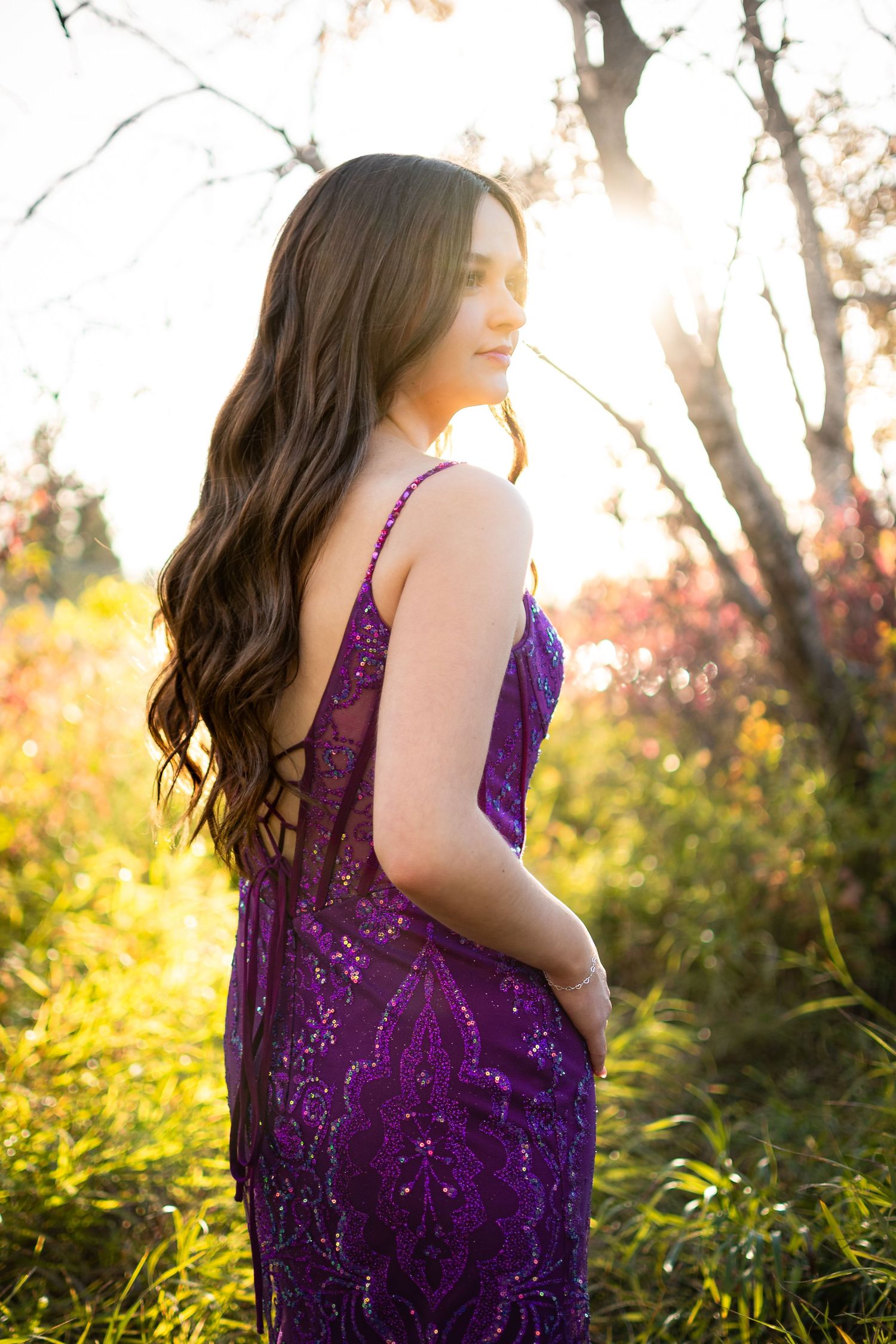 Person in a purple sequined dress posing in natural sunlit setting among trees.