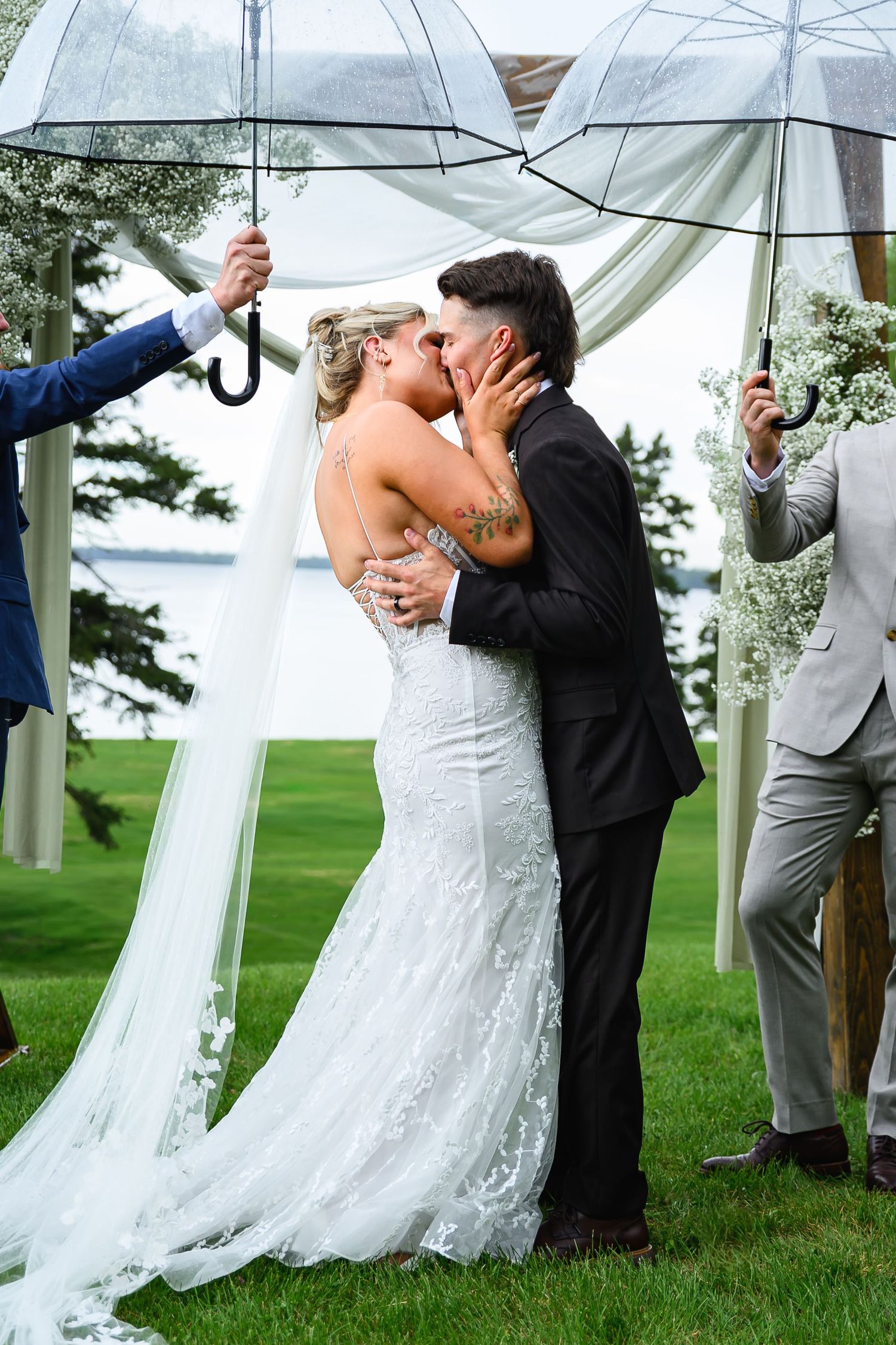 Bride and groom share first kiss under clear umbrellas at outdoor wedding ceremony with white draping and floral arch.