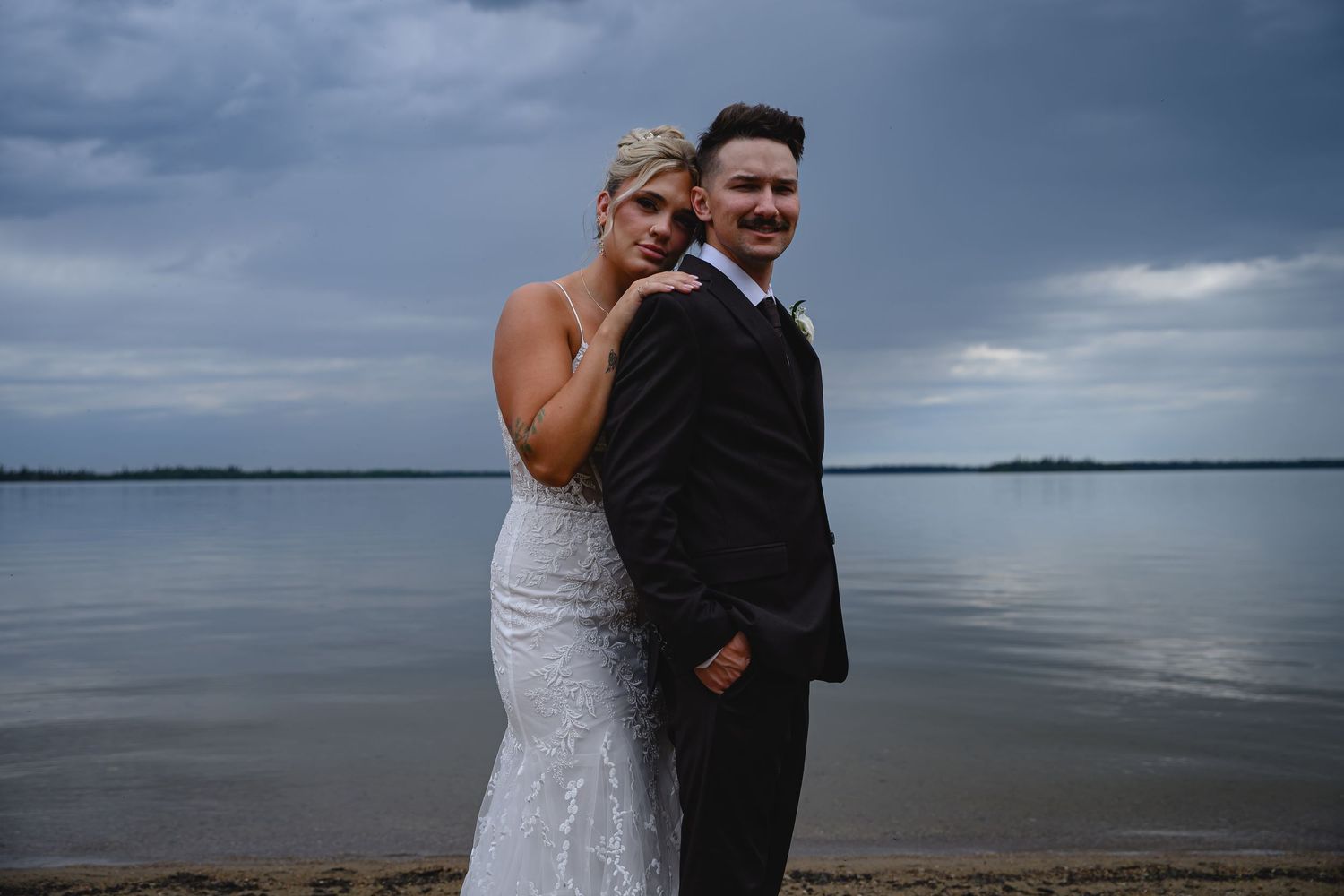 Bride and groom pose romantically on a lakeside beach under dramatic storm clouds in a wedding portrait.