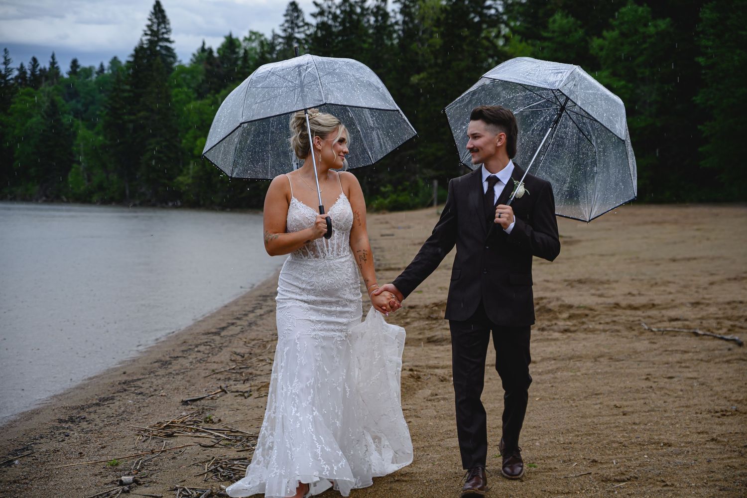 Bride and groom holding clear umbrellas walking hand in hand on a sandy beach on a rainy wedding day.