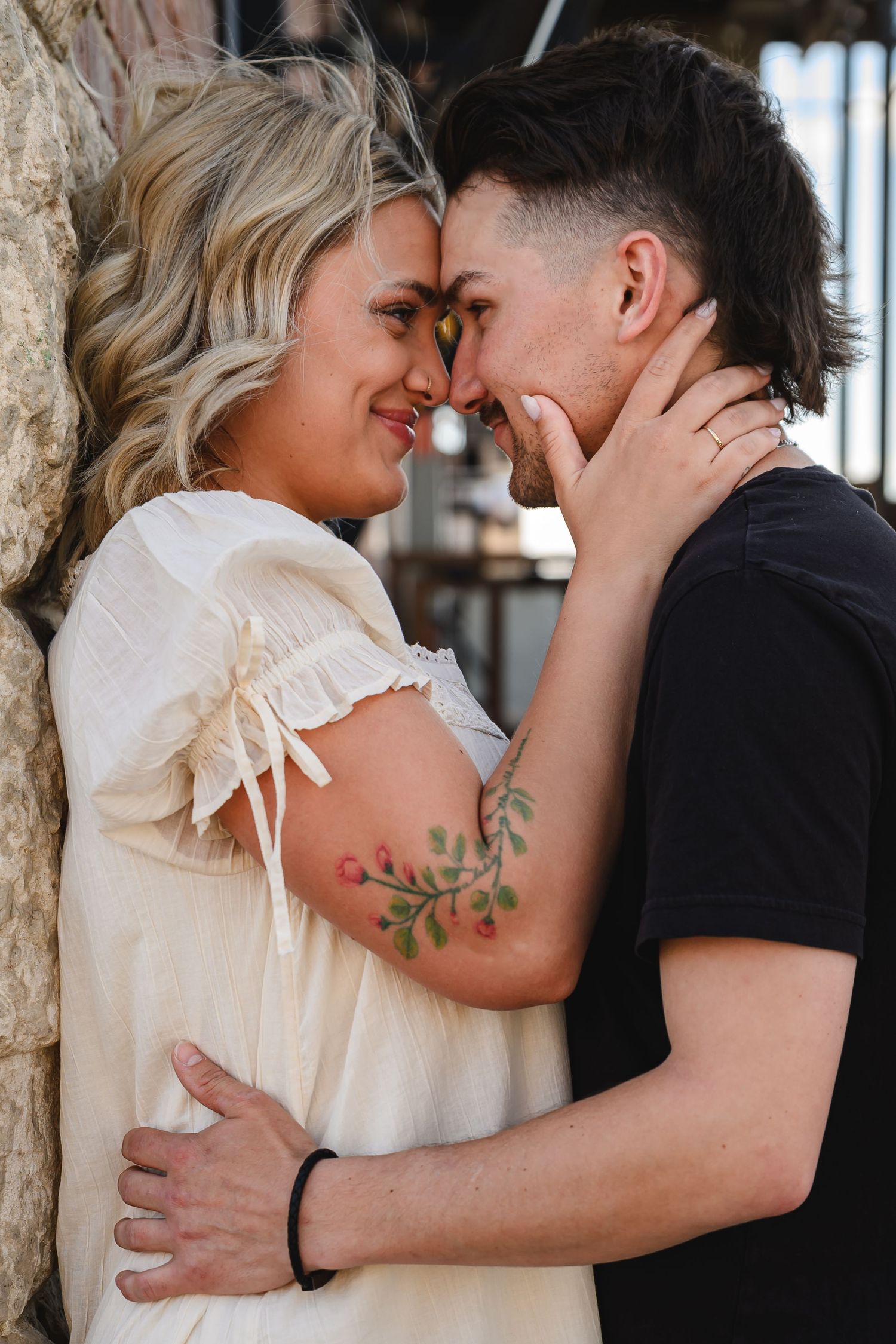 Couple sharing a romantic moment, touching foreheads, woman in white dress with floral tattoo, man in black shirt.
