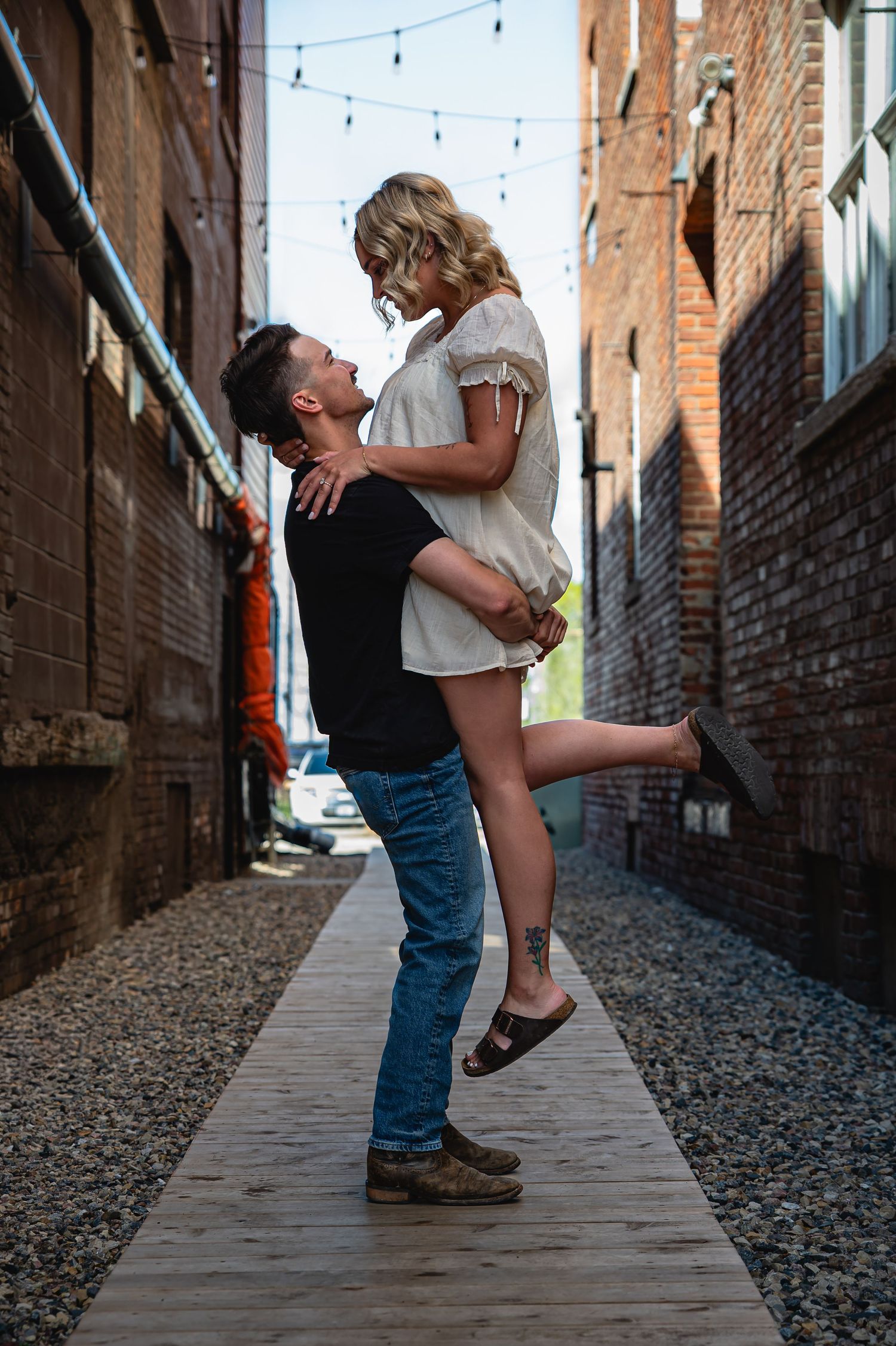 A man lifts a woman in a white dress in a brick alley, sharing a romantic moment outdoors.