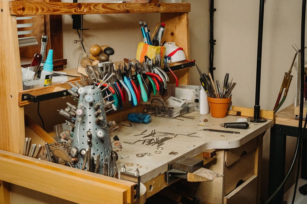 Craftsman working on detailed metalwork at a cluttered workbench with tools and equipment.