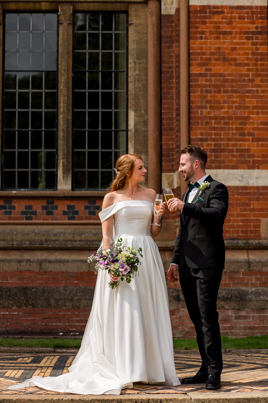 A couple shares laughter while holding drinks during their wedding portraits by a brick building.