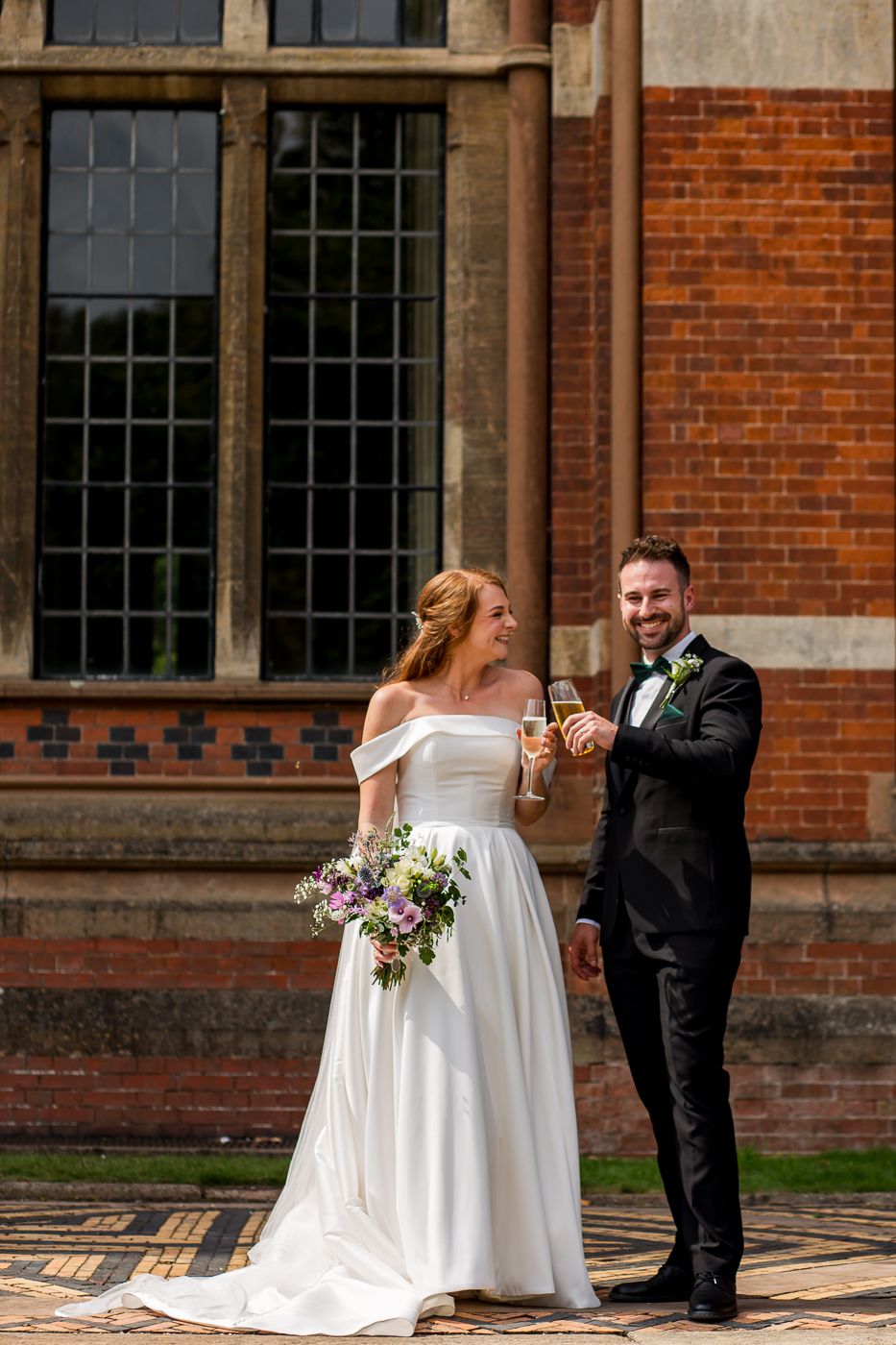A wedding couple toasts with drinks outside a historic brick building with elegant architecture.