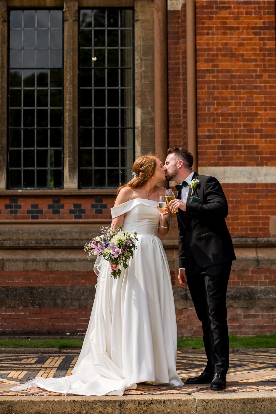 A couple shares an intimate moment while holding drinks against a classic brick building backdrop.