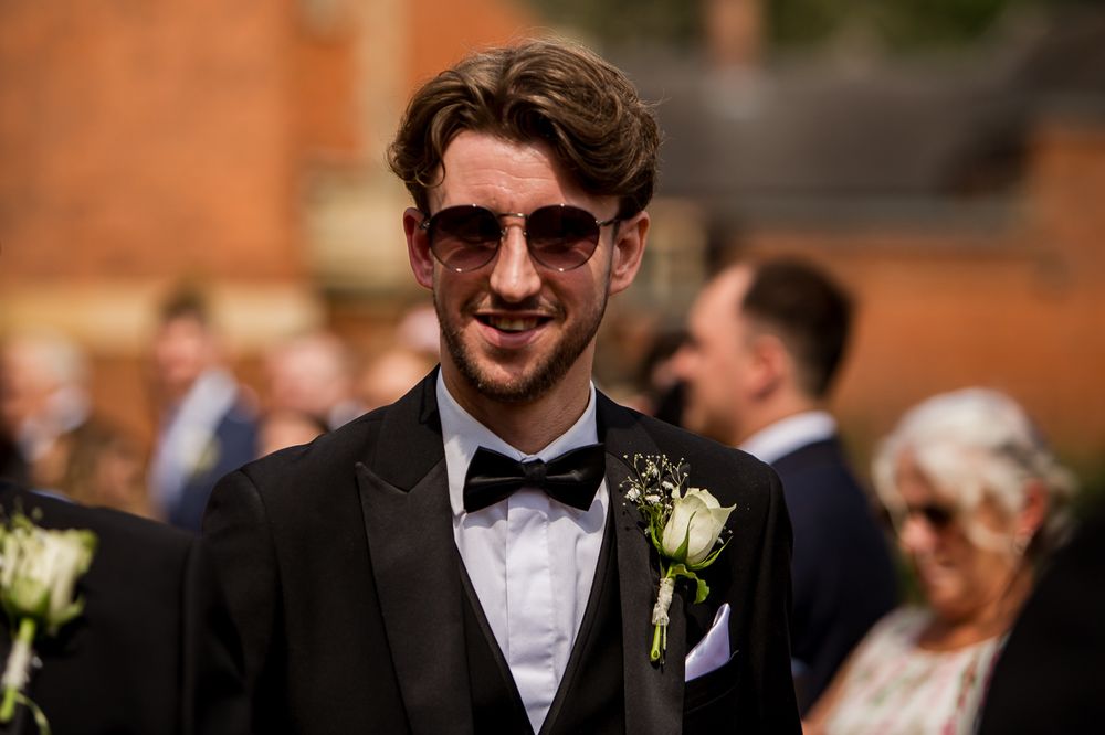 A wedding guest in a black tuxedo and sunglasses smiles during the outdoor celebration.