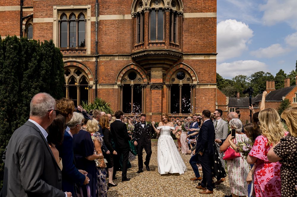 Wedding guests gather outside a grand Victorian Gothic brick building with ornate architecture on a sunny day.
