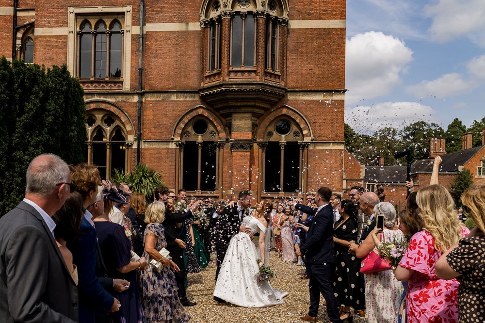 Wedding celebration outside historic red brick building with guests throwing confetti.