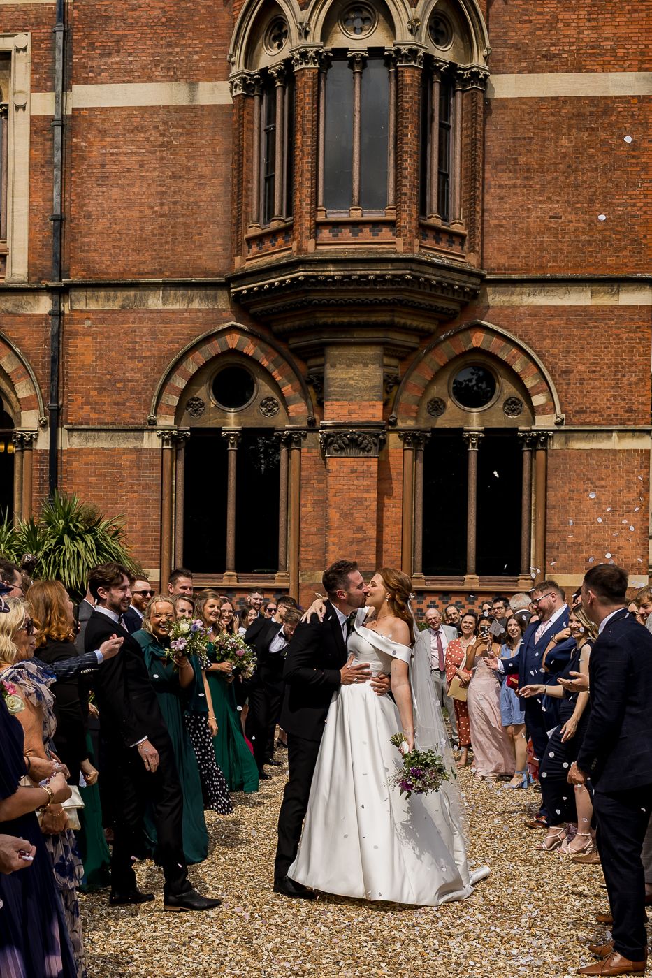 Wedding party and guests celebrate in front of Gothic-style architecture with red brick facade.
