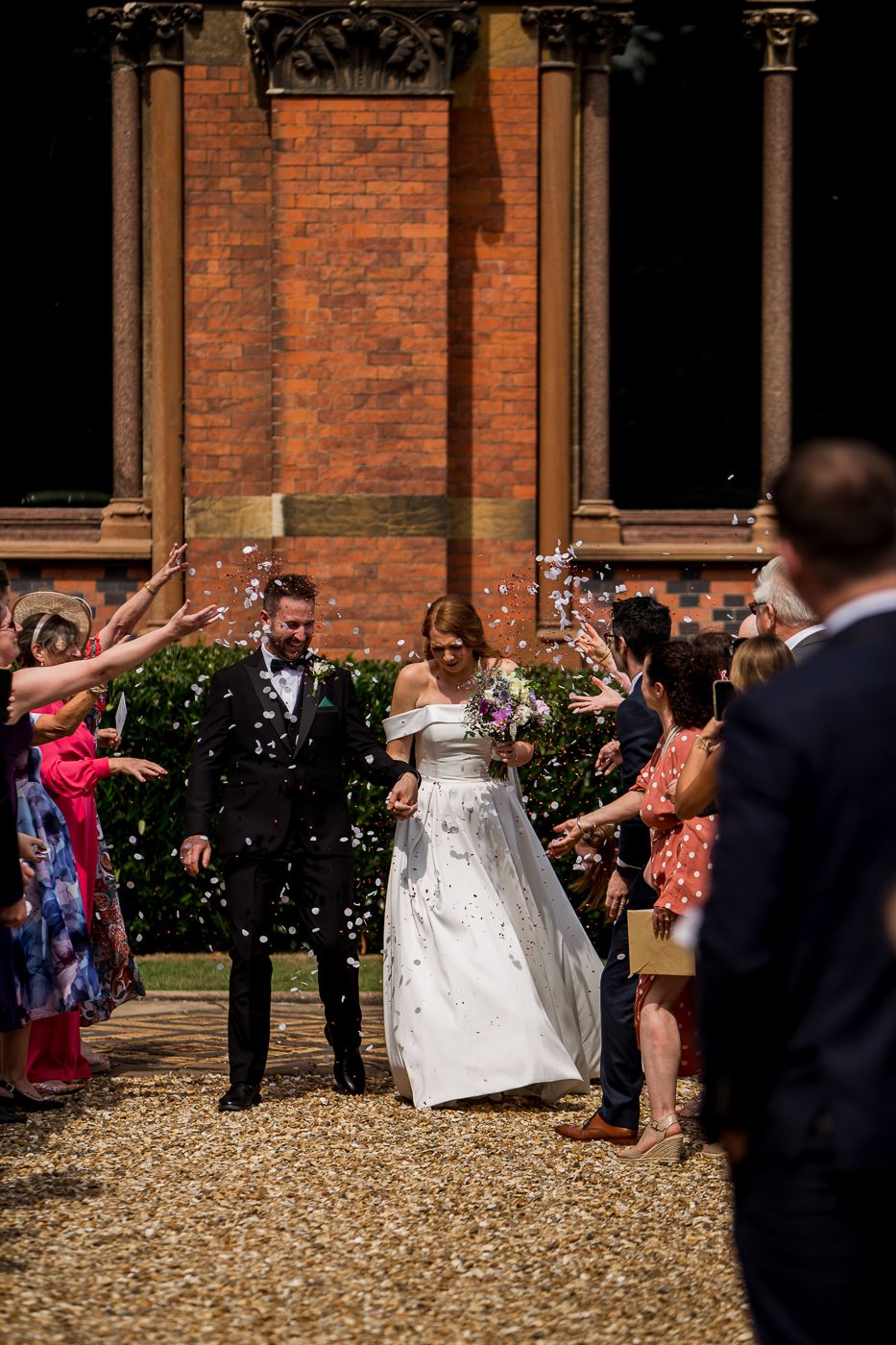 Newlyweds celebrate with guests throwing confetti outside ornate Victorian building.