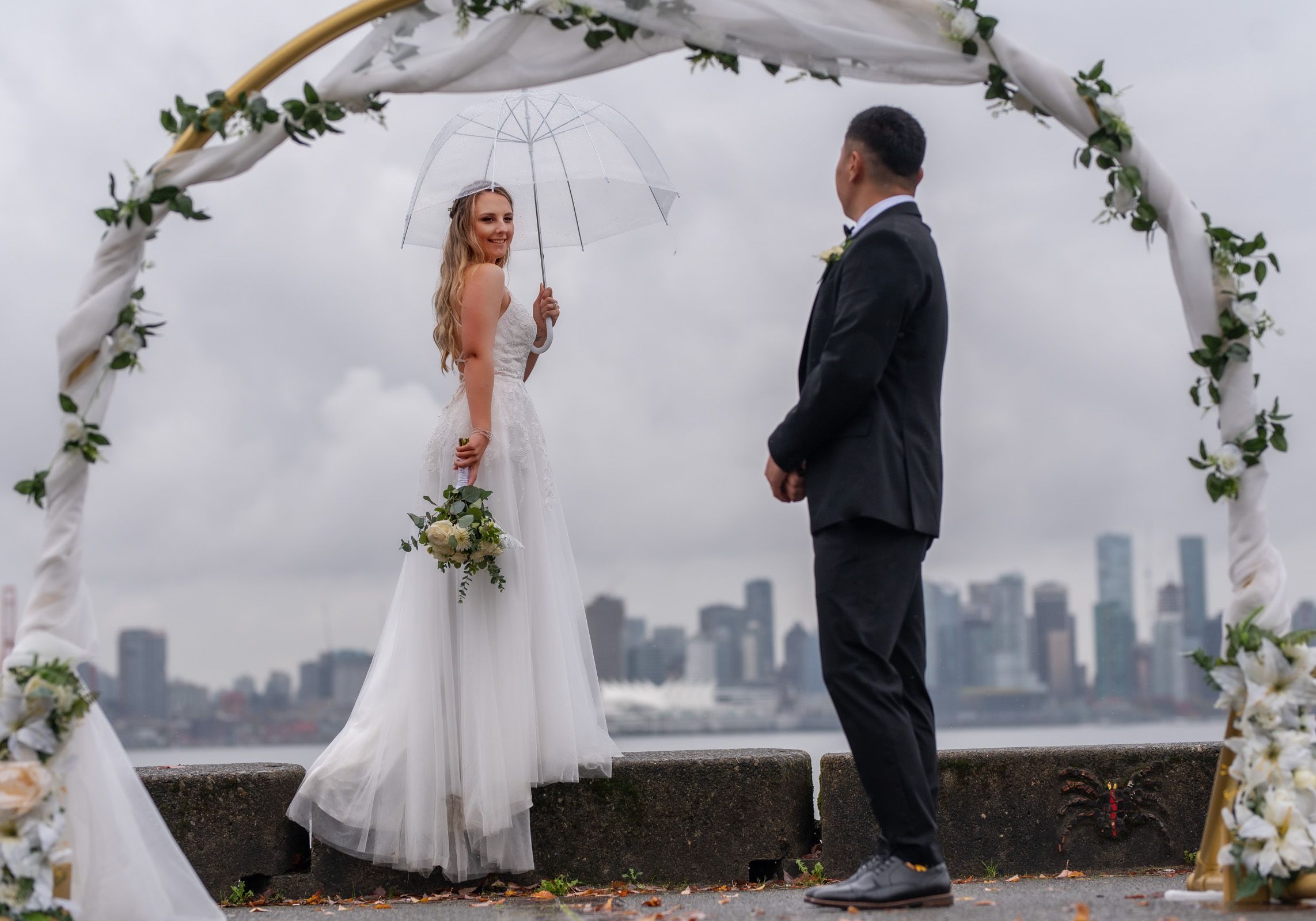 Wedding ceremony under a floral arch with city skyline backdrop.