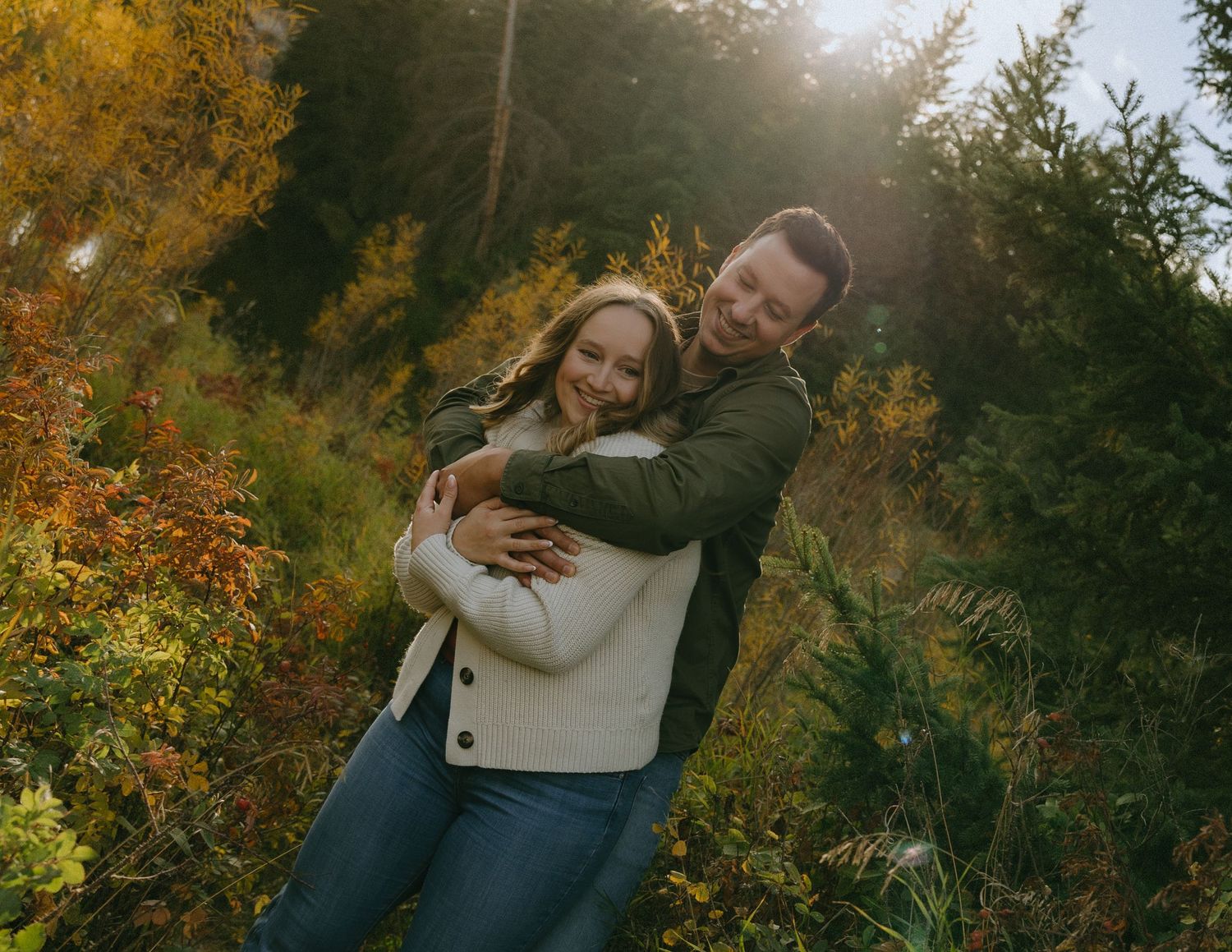 A couple embraces outdoors surrounded by golden autumn foliage and sunlight filtering through trees.