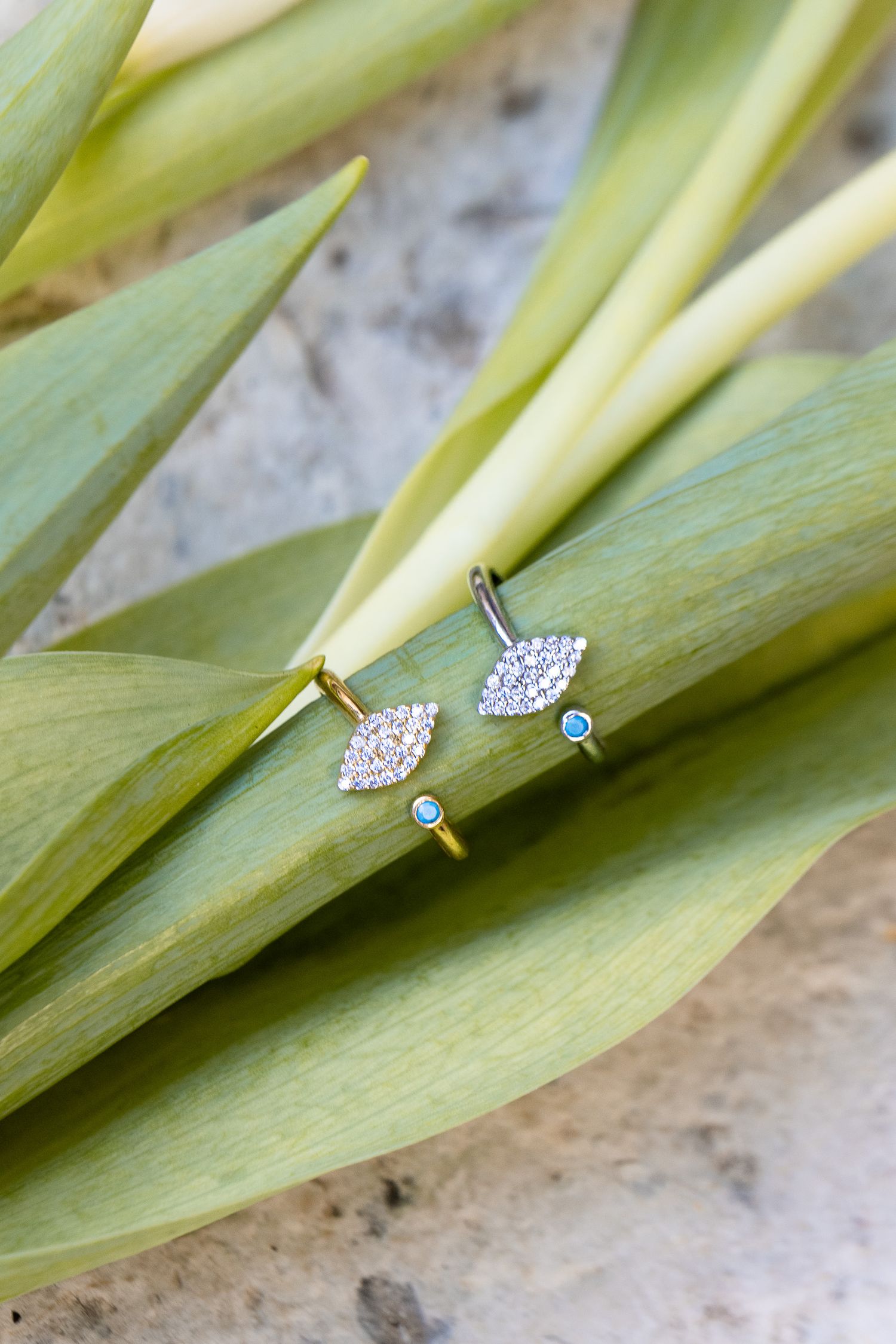 Close-up of delicate diamond and sapphire stud earrings displayed on fresh green leaves against a stone background.