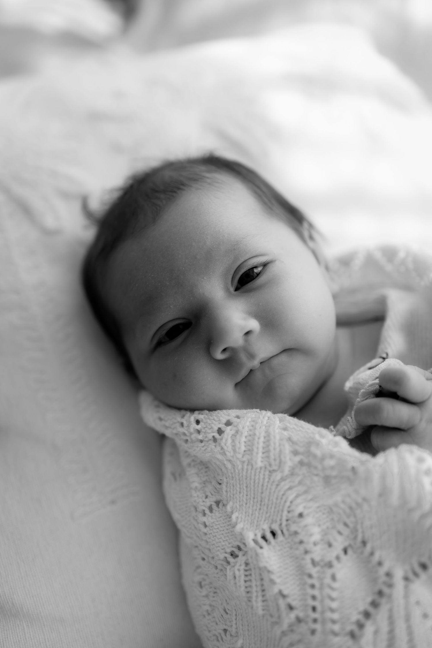 Sweet newborn baby wearing delicate white lace lies peacefully against soft white bedding.