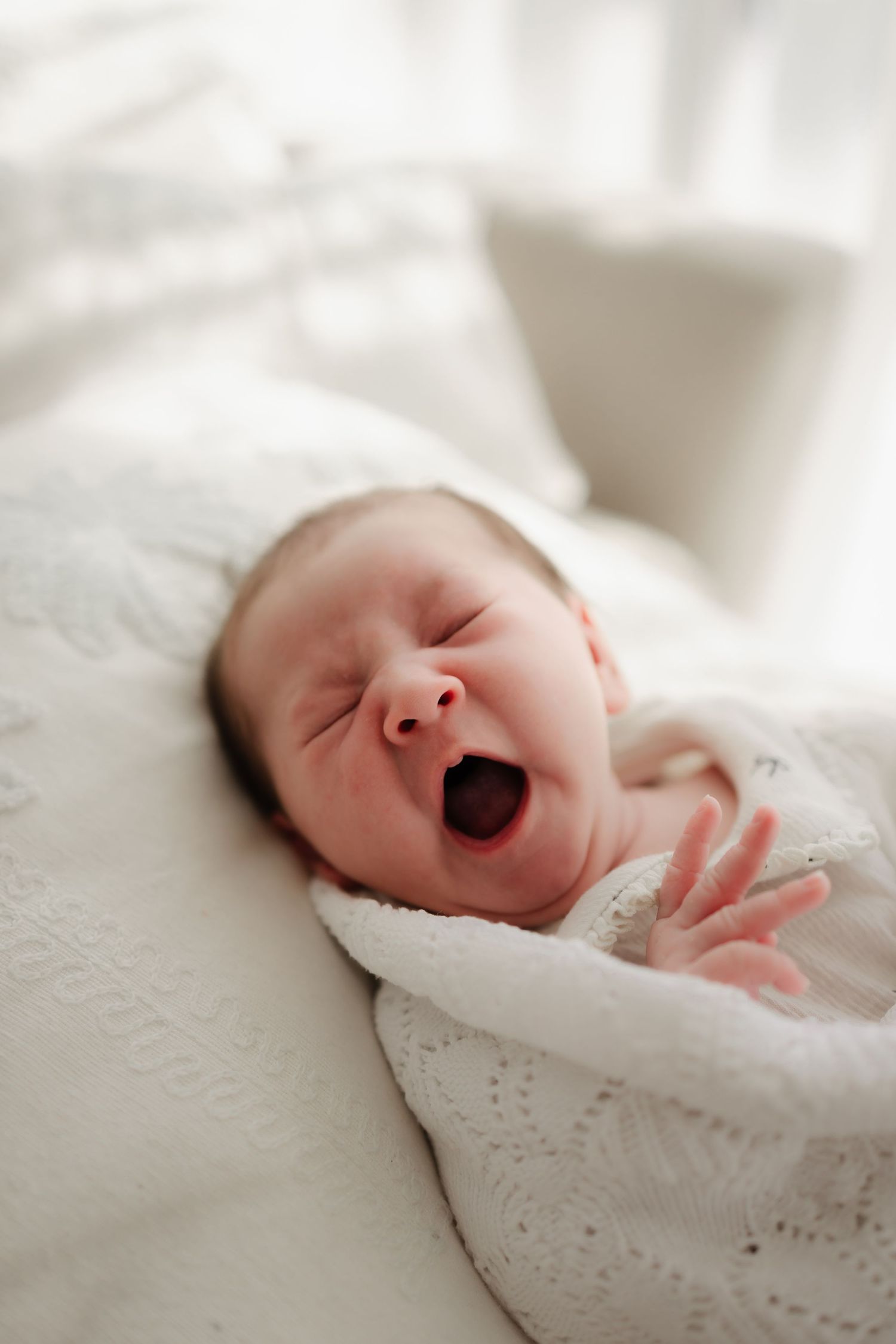 Sleeping newborn baby in white lace yawns while resting on white blankets.
