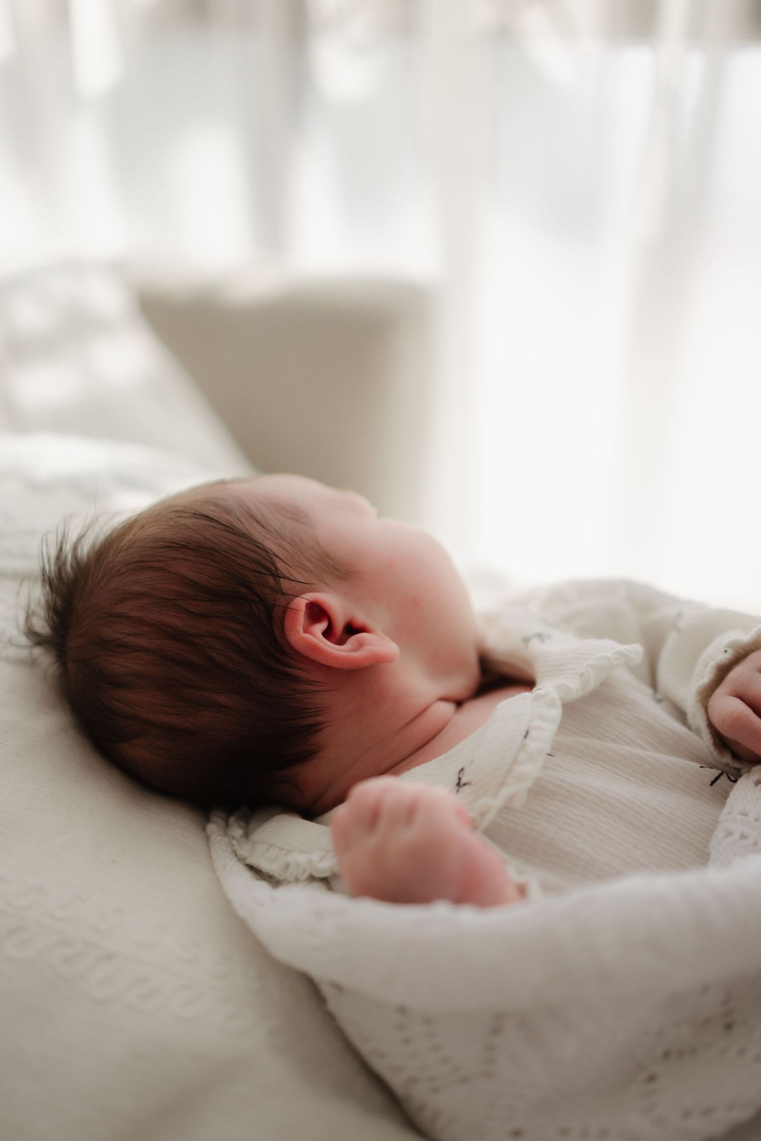 A sleeping newborn baby rests peacefully on soft white bedding in natural light.